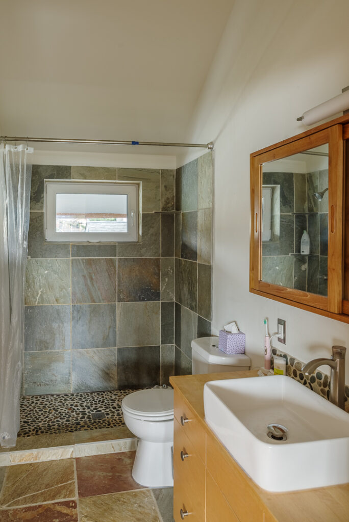 A photo of the bathroom looking north. There is a single sink vanity, toilet and walk-in shower with a small operable window. 