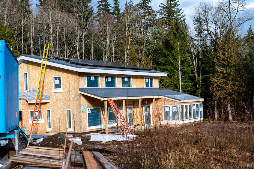 A photo of the exterior of a home under construction. The homes framing is completed, windows and roofing are already installed, and all but a few solar panels have been installed, but it's clear the photo was taken while the solar installers were in the process of installing. This home has a big covered porch over the south side first floor and an overall clearstory design.