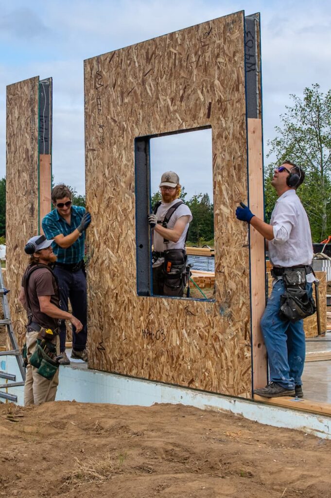 A photo of 3 crew members placing a wall panel with a window opening on the slab. 1 crew member is watching and guiding. There is a piece of wood seen nestled in the edge of the panel.