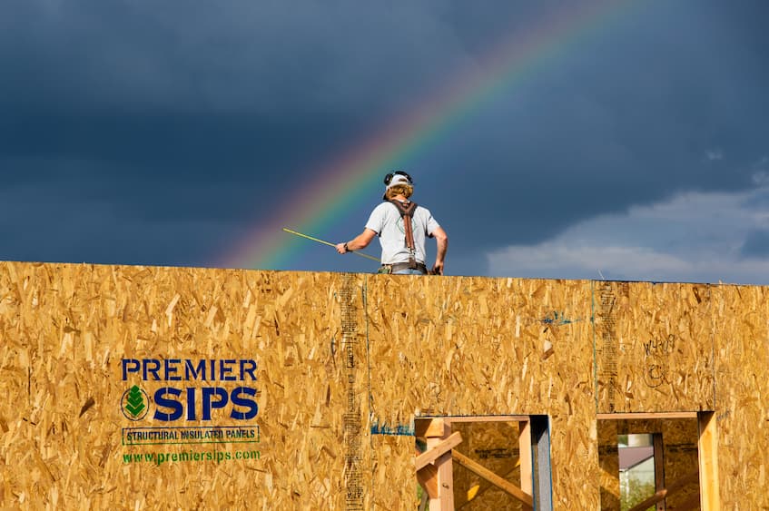 What are SIPS? This is a photo of owner Norm back to the camera, holding a tape measurer on top of an under construction home. The walls below him are SIPs and labeled "Premier SIPs Structural Insulated Panels www.PremierSIPs.com". There is a rainbow just above Norm's head and the sky is dark.