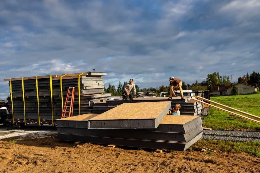 A photo of 4 crew members off loading a large stack of SIPs from a truck trailer. Two men stand on top of the panels on the truck, and two on the ground guiding the panels onto the ground. The panels appear to be at least 5' wide by 10' tall. You can see the layers of the panels (wood sandwiching foam) with 2 holes running the entire height for wires.