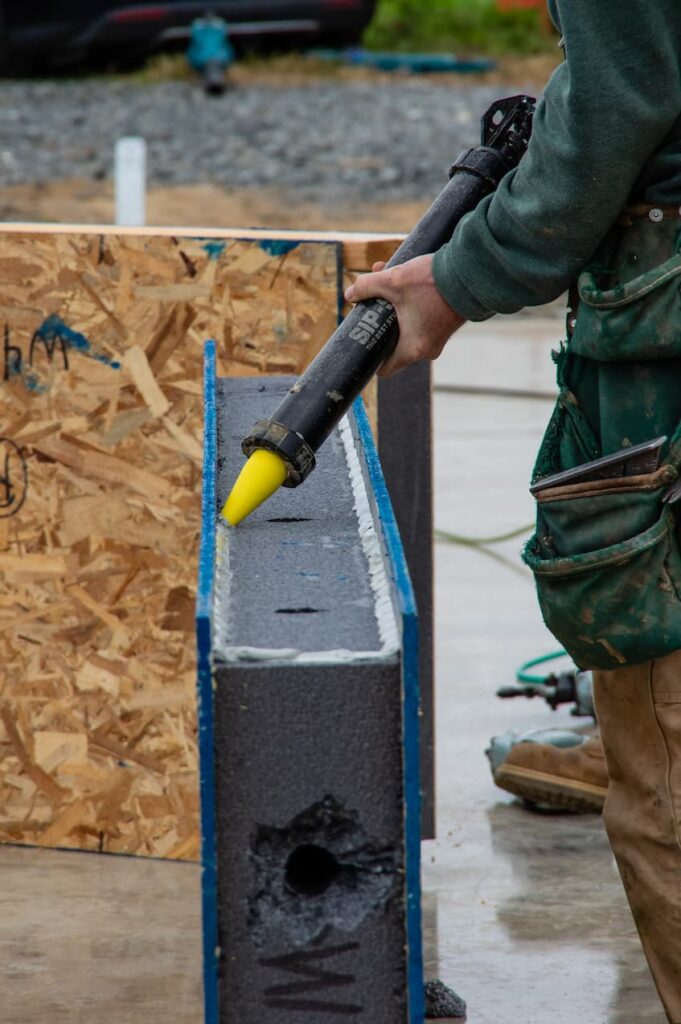 A close-up photo of a man applying SIP glue through a large caulking gun directly to the foam along the edges of the panel, following the entire perimeter.