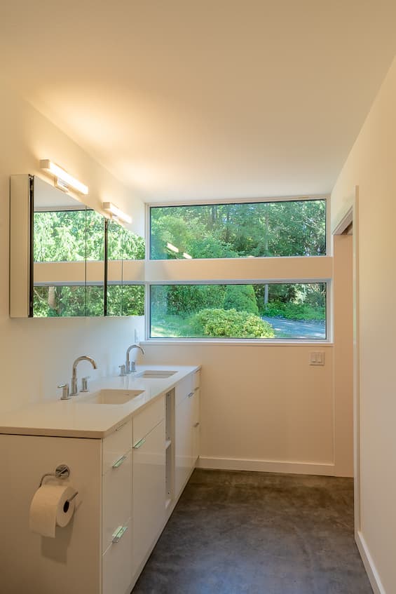 Photo inside a bathroom looking towards the double sink vanity and large windows. The vanity, countertops and walls are all white. The green of the nature outside is bouncing off the mirrors above the sink.