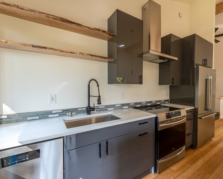 A photo inside a kitchen looking at the sink, range and fridge. The cabinets are glossy gray and the appliances are stainless steel. The range has an induction glass stove top.