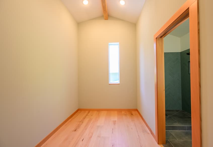 A photo of an empty room with vaulted ceilings, warm wood floors, warm wood trim. There is a tall and skinny window on the back wall with a warm wood sill. And a door on the right is cased in warm wood trim.
